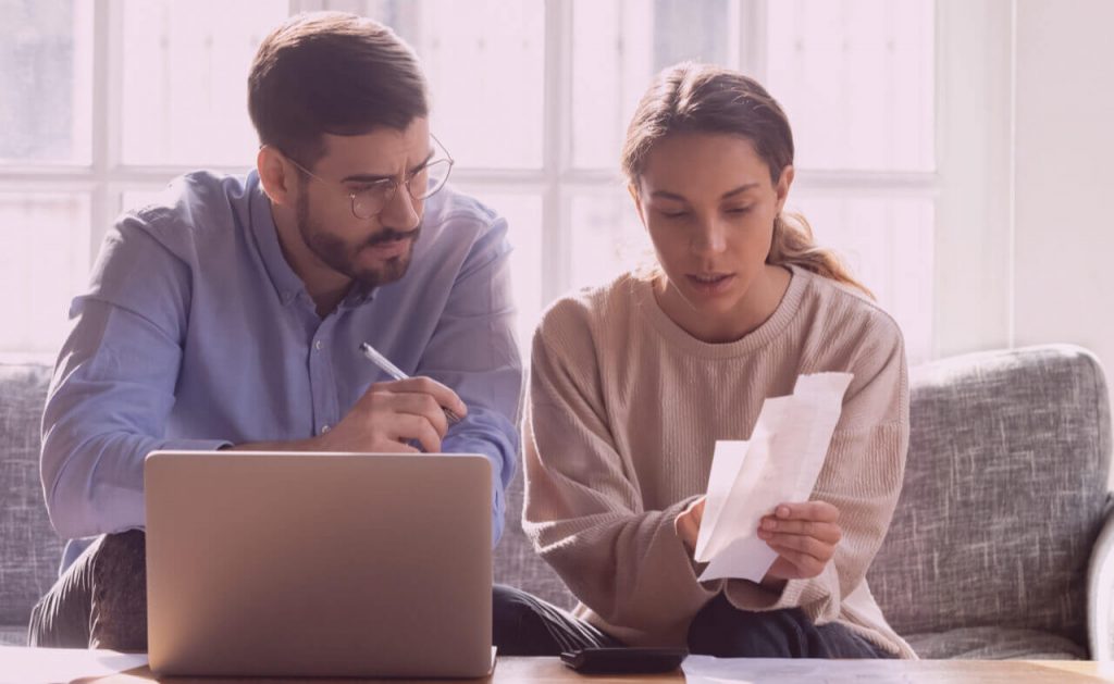 Serious couple calculating expenses, bills together, using laptop, sitting on couch at home.